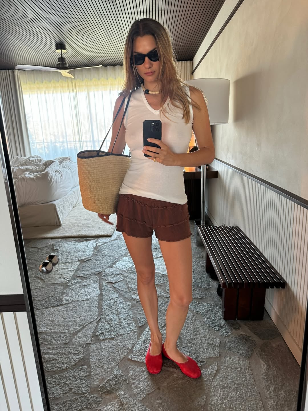 Μια κοπέλα φοράει ένα μίνιμαλ σύνολο με λευκή αμάνικη μπλούζα με V λαιμόκοψη, χαλαρό καφέ λινό σορτς και έντονες κόκκινες φλατ μπαλαρίνες που δίνουν ένα pop χρώματος. Στον ώμο της κρατάει μια μεγάλη ψάθινη τσάντα-καλάθι με δερμάτινα λουράκια.