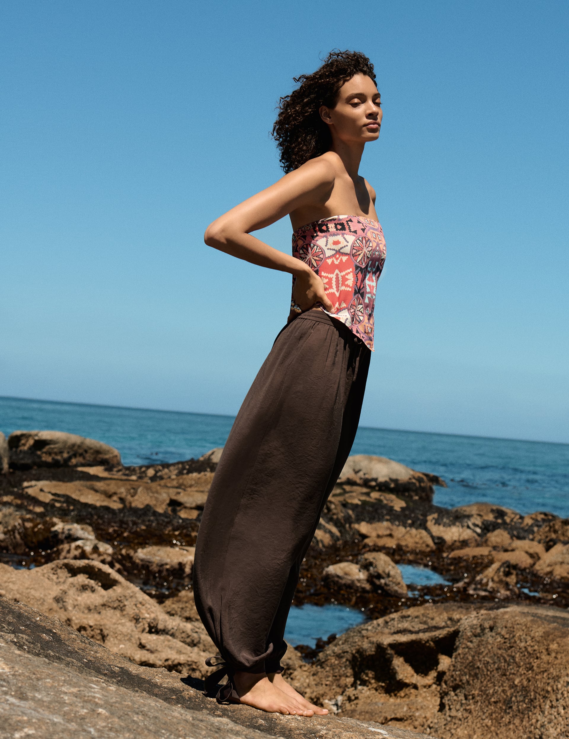 Γυναίκα μοντέλο φοράει printed top Marks & Spencer με καλοκαιρινά μοτίβα, ποζάροντας με φόντο τη θάλασσα.