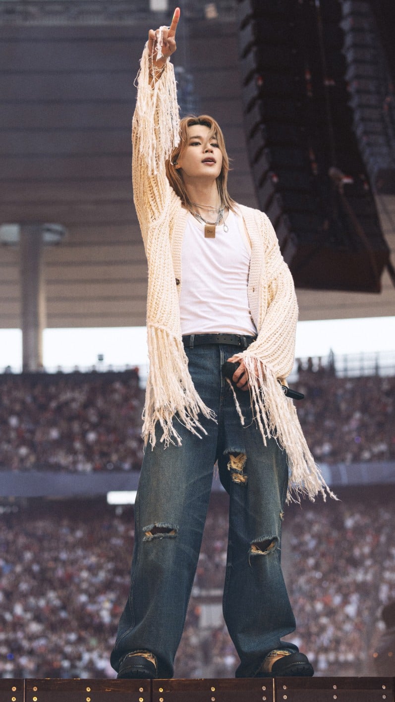 Ο Jimin με Dior σε ένα relaxed look, συνδυάζοντας off-white μάλλινο knit με λευκό βαμβακερό tank top και μπλε denim jeans. Ολοκλήρωσε την εμφάνισή του με μαύρη δερμάτινη ζώνη και μπεζ δερμάτινα lace-up boots.