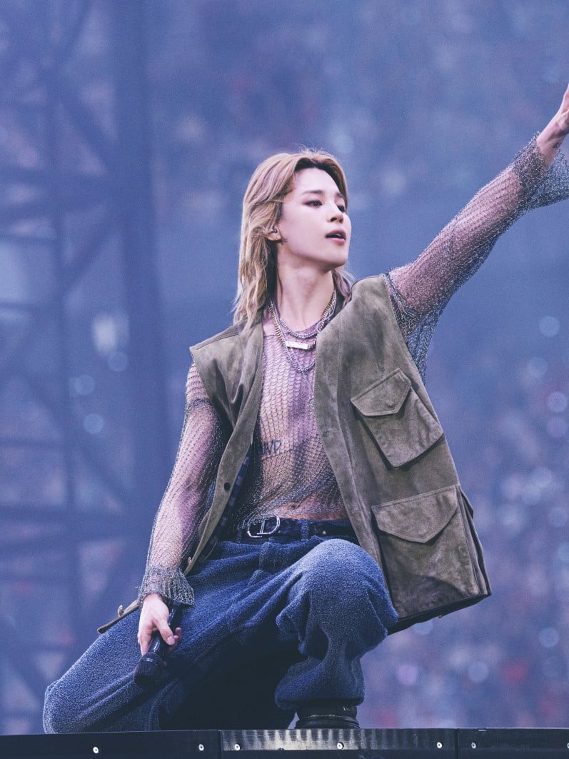 Ο Jimin με ένα statement Dior look, συνδυάζοντας khaki αμάνικο δερμάτινο jacket με silver lurex πλεκτό top και navy washed cotton παντελόνι σε φαρδιά γραμμή. Ολοκλήρωσε την εμφάνισή του με μαύρη braided leather ζώνη και μαύρα δερμάτινα παπούτσια.